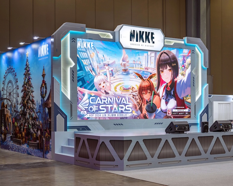 A futuristic-themed exhibition booth for the subculture game "NIKKE: Goddess of Victory" at AGF 2024. The booth features a large LED screen displaying the event title "Carnival of Stars" with anime-style characters, a performance stage with speakers, and immersive wall graphics of a festive theme park with a Christmas tree and ferris wheel.